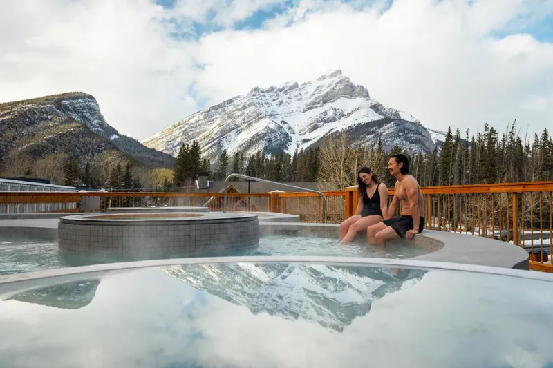 Couple sit in outdoor hot pool with a snowy mountain backdrop